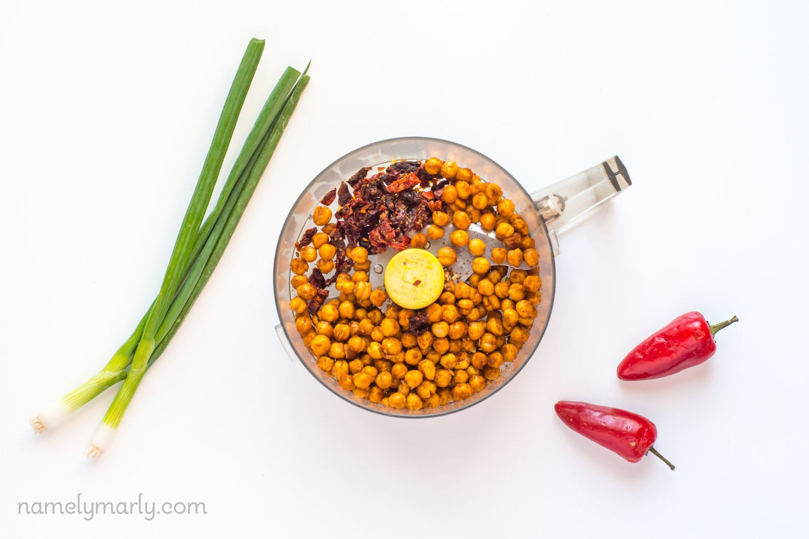 Looking down on a food processor full of sun-dried tomatoes and roasted chickpeas. Green onions and mini red bell peppers sit next to it.
