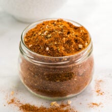 A small glass jar is full of homemade taco seasoning sits in front of a bowl with more of the seasoning.