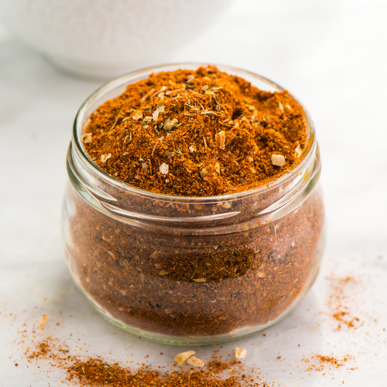 A small glass jar is full of homemade taco seasoning sits in front of a bowl with more of the seasoning.