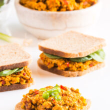 A piece of bread is topped with vegan deviled ham spread. Two other sandwiches sit behind it, along with a bowl of more spread.