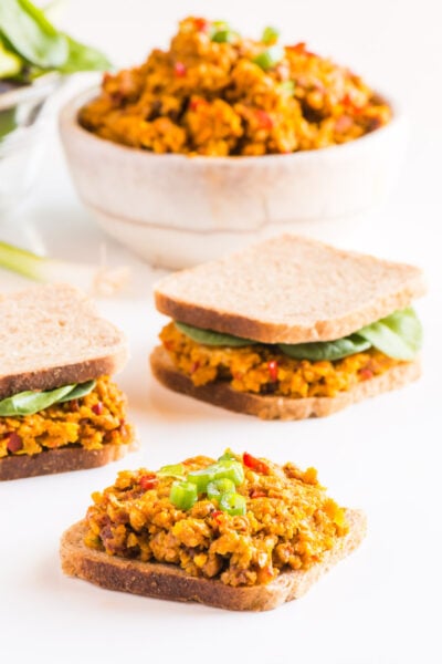 A piece of bread is topped with vegan deviled ham spread. Two other sandwiches sit behind it, along with a bowl of more spread.