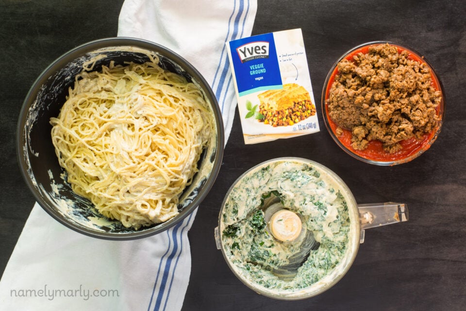 A bowl of cooked pasta sits on a white kitchen towel. Next to it is a food processor bowl with the tofu and spinach already pulsed. Next to these is a plate with Yves veggie crumbles and the container it came in.