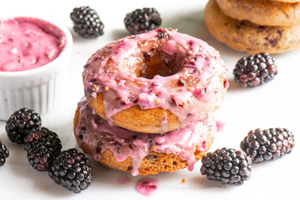 Two glazed blackberry donuts surrounded by fresh blackberries and a bowl of blackberry glaze.