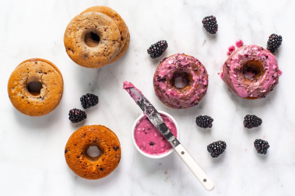Looking down on two glazed vegan blackberries donuts sitting next to blackberries and a bowl of blackberry glaze and several unglazed donuts.
