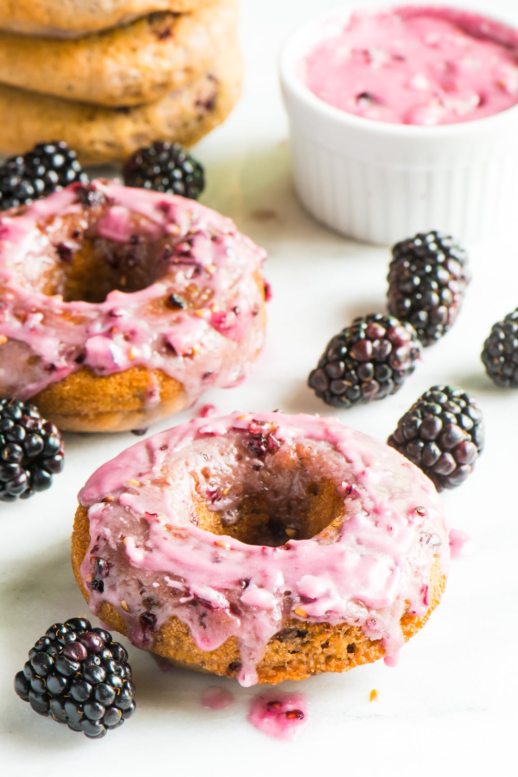 Two vegan blackberry donuts sit next to blackberries. A bowl of blackberry glaze and a stack of unglazed donuts sits behind it.