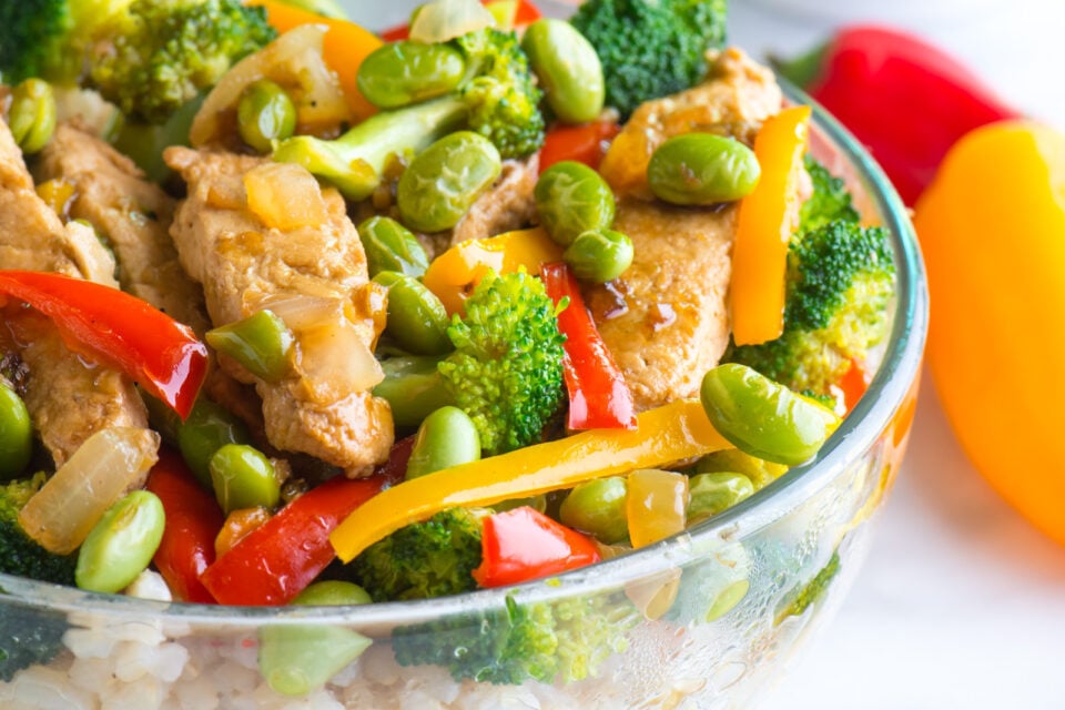 A closeup of a glass bowl full of vegan teriyaki chicken over brown rice and topped with edamame. Two mini bell peppers are beside it. 