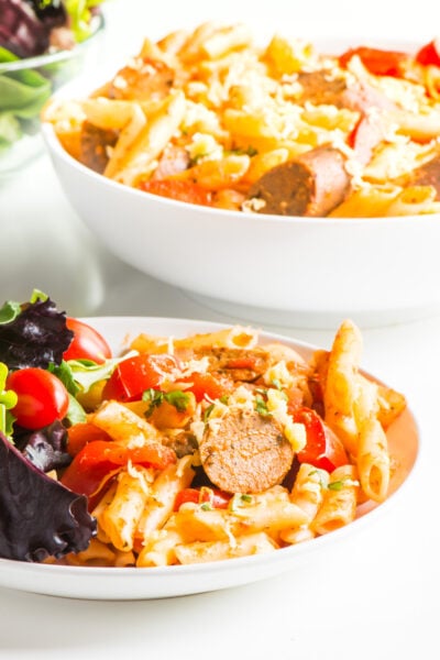 Two pasta bowls full of easy vegan penne pasta sit one in front of the other with salads on the side.