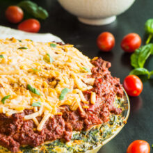 A pie plate full of vegan spaghetti pie with melted vegan cheese on top sits on a dark countertop. There are fresh cherry tomatoes sitting around the the dish, with fresh basil and a bowl full of pasta sauce in the background.