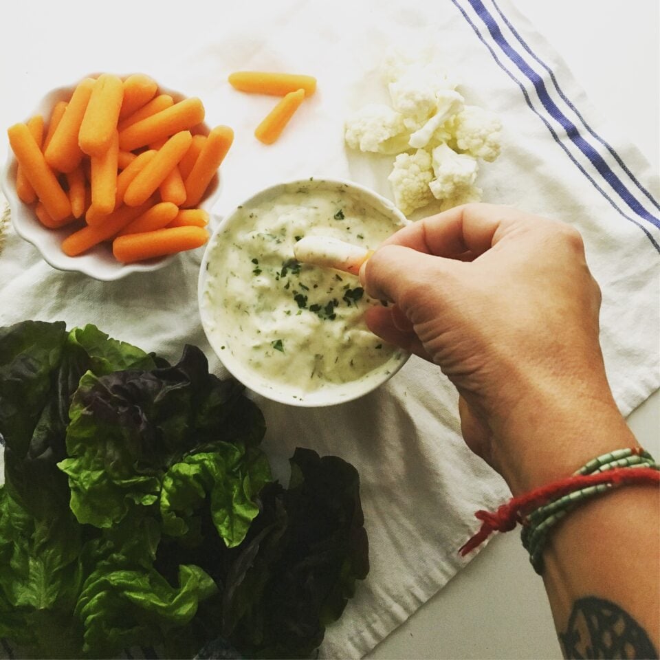 Looking down on a bowl of vegan ranch dip with a hand reaching in to dip a baby carrot.