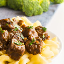 A white plate topped with vegan beef stroganoff shows noodles and parsley sprinkled over the vegan beef pieces. There is steamed broccoli in the background.