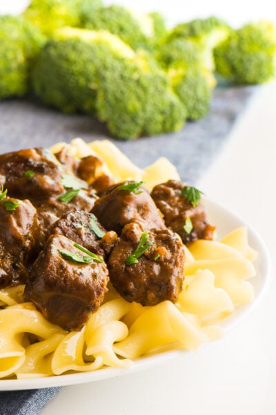 A white plate topped with vegan beef stroganoff shows noodles and parsley sprinkled over the vegan beef pieces. There is steamed broccoli in the background.