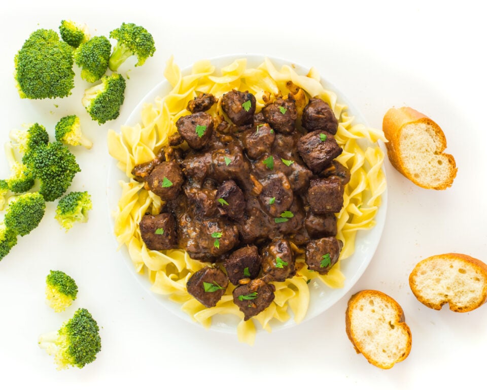 Looking down on a plate of vegan beef stroganoff, surrounded by slices of bread and steamed broccoli florets.