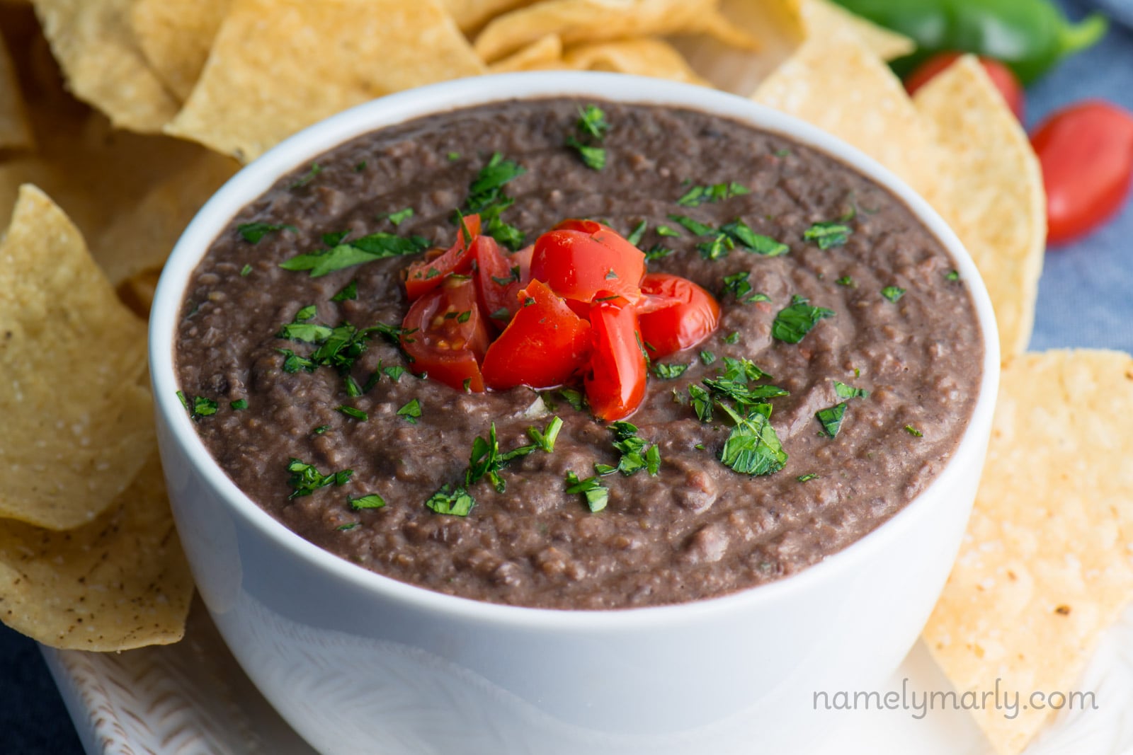 A bowl of Mexican spread with chopped tomatoes on top sits next to some tortilla chips.