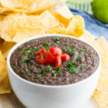 A bowl of bean dip next to tortilla chips with a lime and a margarita behind it.