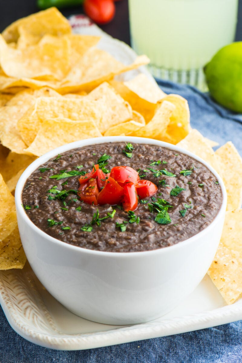 A bowl of bean dip next to tortilla chips with a lime and a margarita behind it.