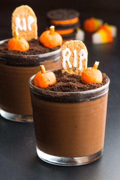 Two small glasses hold chocolate pudding with candy pumpkins and a RIP graham cracker tombstone. These are vegan Halloween Pudding cups to serve at parties.