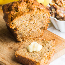 A slice of vegan banana bread with a pat of butter on it sits in front of the rest of the loaf.