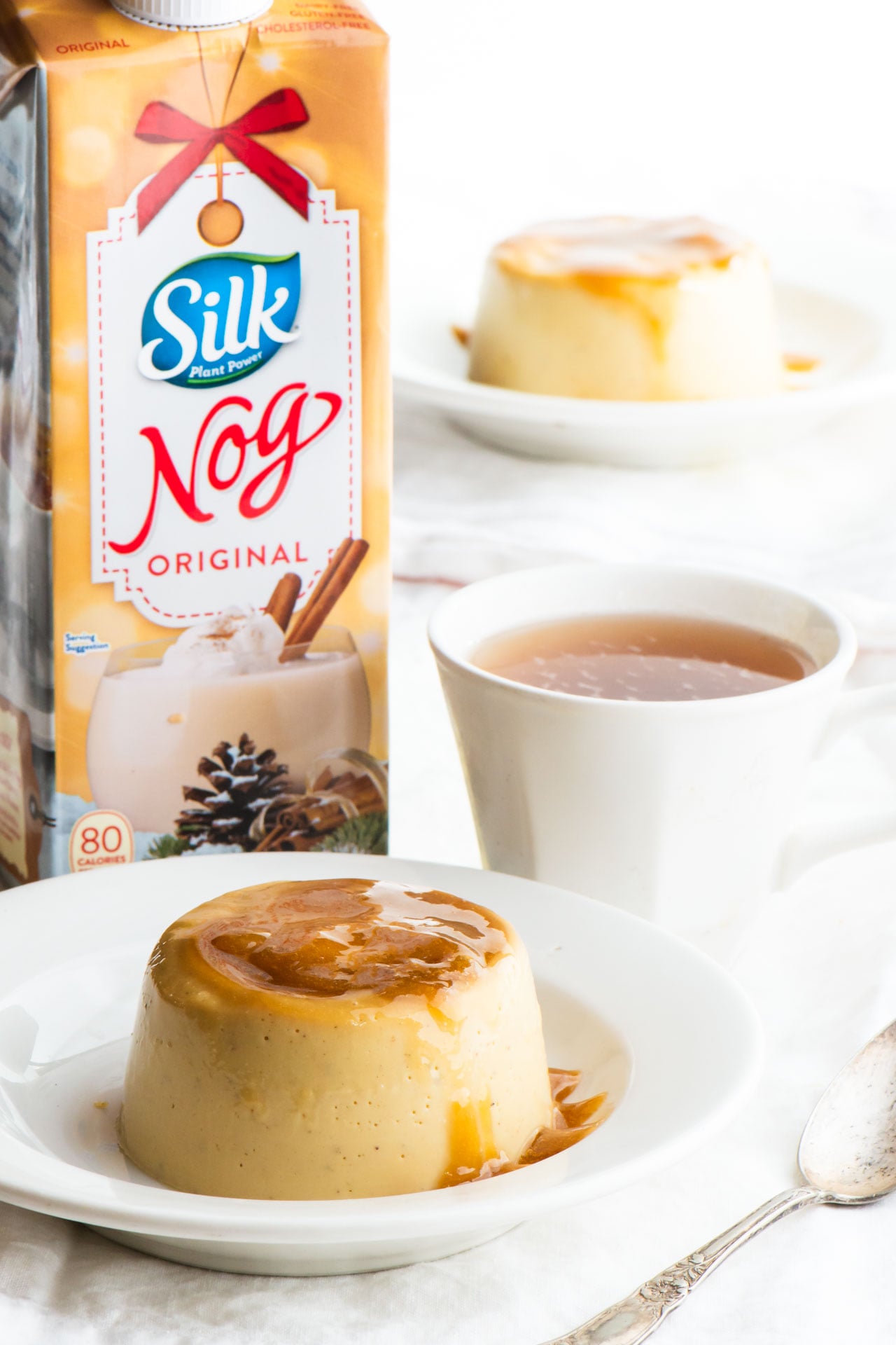 Two plates of vegan flan with a cup of tea and a container of Silk dairy-free Nog.