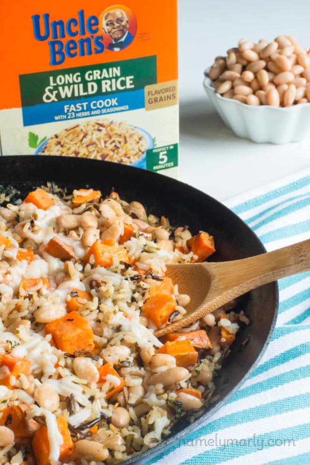 A close-up of a skillet full of wild rice dinner with a box of rice behind it and a bowl of white beans.
