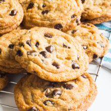 A freshly-baked batch of vegan coconut chocolate chip cookies sit on a wire rack to cool. A bowl of chocolate chips sits behind them.