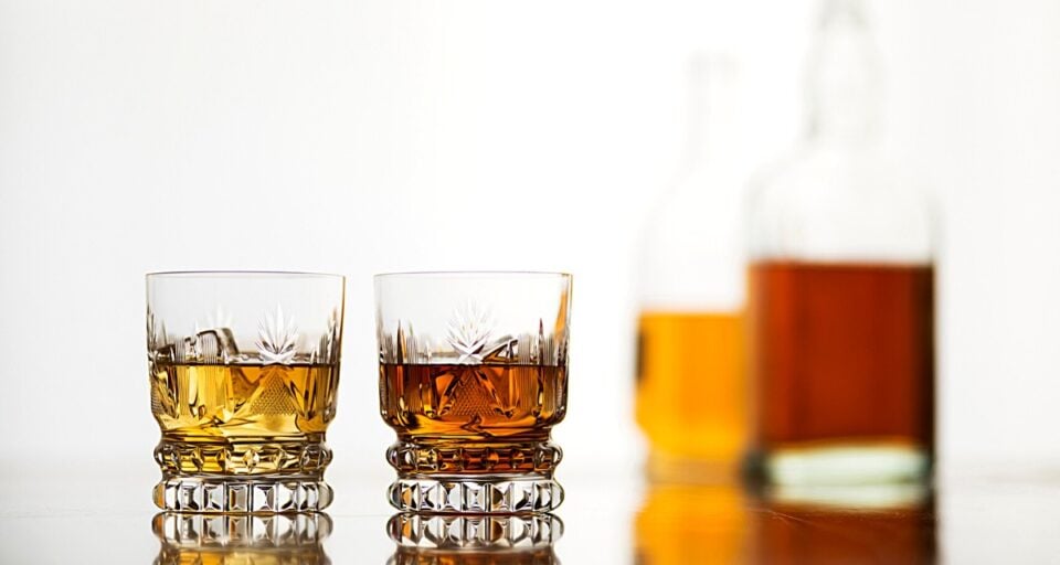 Two glasses of whiskey sit on a reflective counter top. There are two bottles of whiskey in the background.