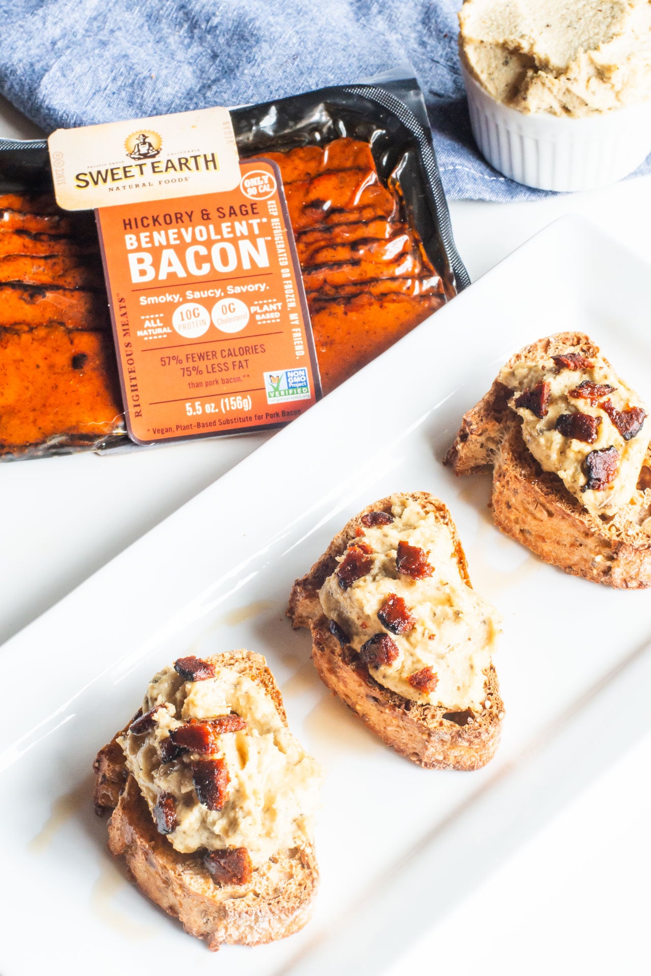 Several slices of vegan crostini are on a plate with a package of vegan bacon behind it.