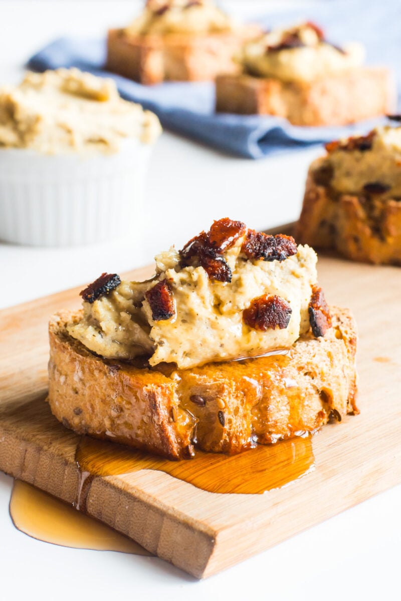 A slice of vegan bacon ricotta crostini with maple syrup drizzling over it on a cutting board.