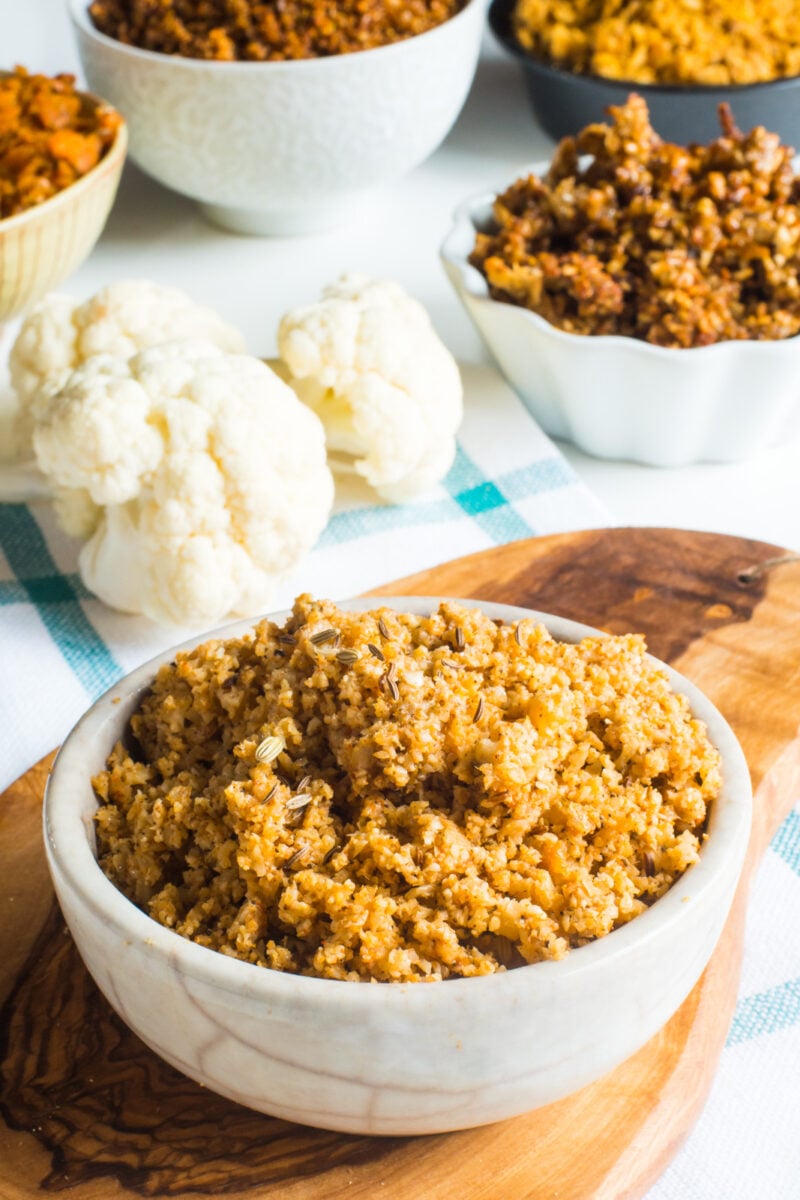 A bowl of Vegan Cauliflower Sausage Crumbles sits on a cutting board in front of raw cauliflower and bowls of other vegan sausage substitutes.