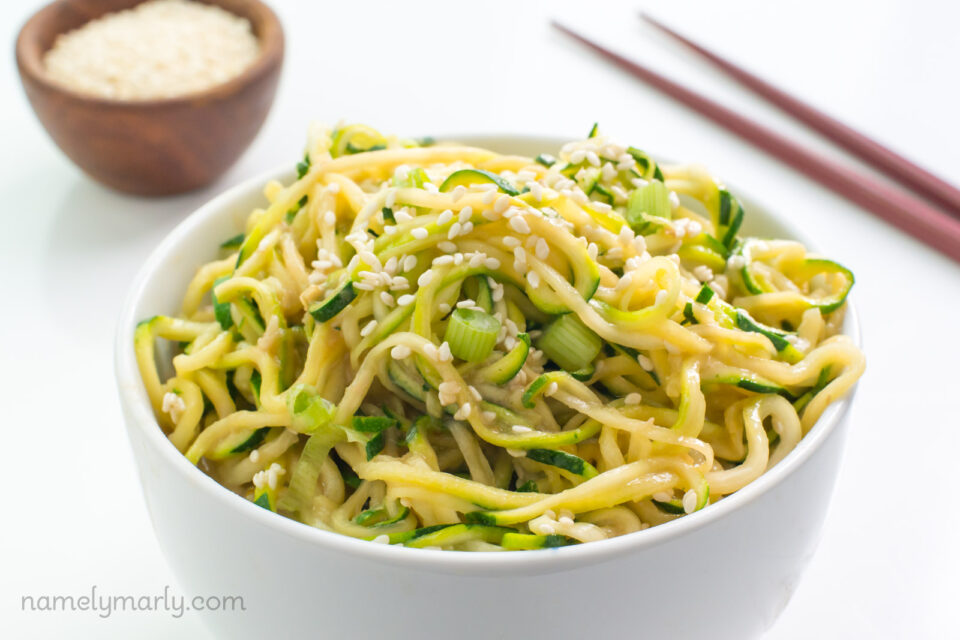 A bowl of spicy sesame zoodles shows chopsticks behind it and a bowl of sesame seeds.