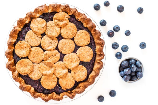 An overhead view of a berry pie with blueberries scattered around on a marble surface