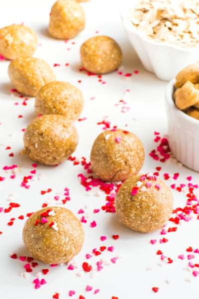 A group of Sugar Cookie Energy Bites sits on a white surface surrounded by red and pink heart-shaped confetti