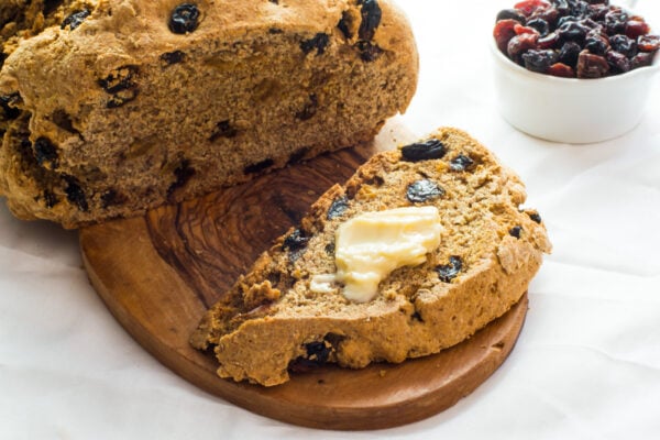 A loaf with raisins has a slice cut out with melted vegan butter and a bowl of raisins beside it.