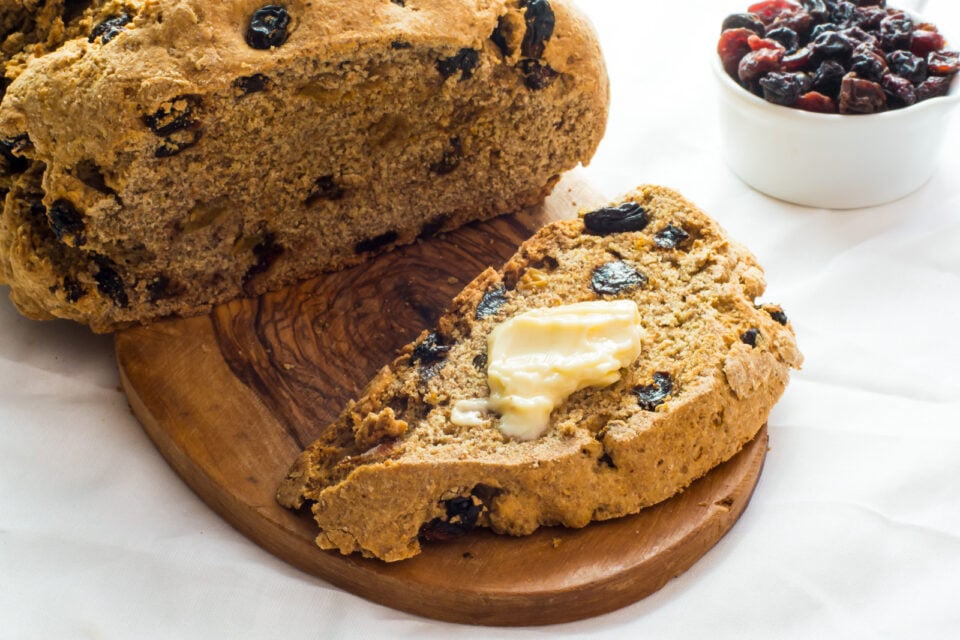 A loaf with raisins has a slice cut out with melted vegan butter and a bowl of raisins beside it.