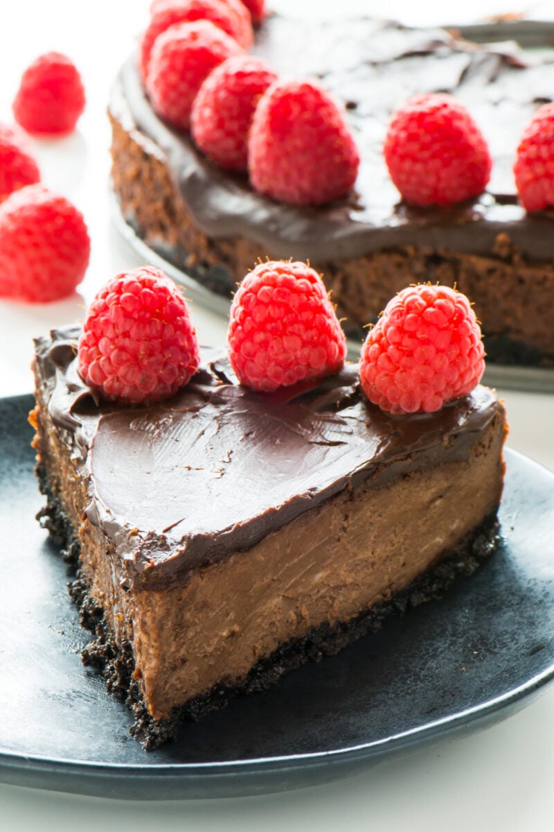 A slice of vegan chocolate cheesecake has a row of fresh raspberries on top. The rest of the cheesecake is sitting behind the slice.