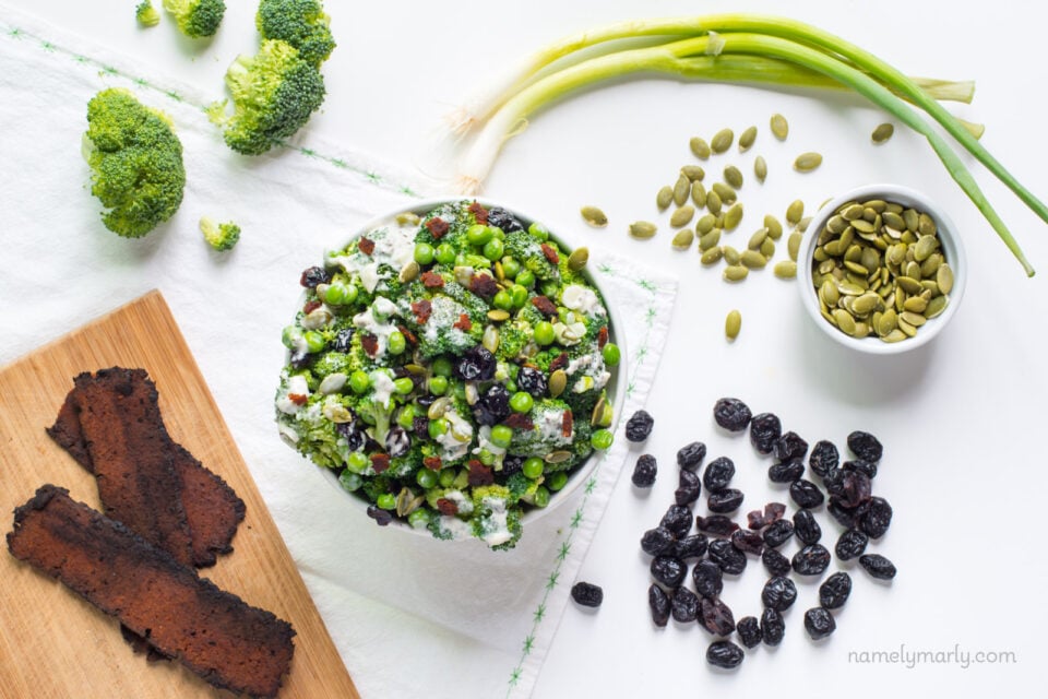 Vegan Broccoli Salad sits in the middle of a table, a bowl of pumpkin seeds sits on the side, with raisins nearby and some green onions. There are more broccoli pieces on the side and some pieces of veggie bacon on a cutting board.