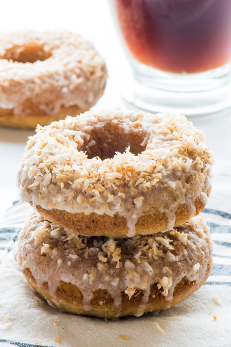 A stack of toasted coconut donuts sits on a kitchen towel with another donut in the background next to a cup of tea
