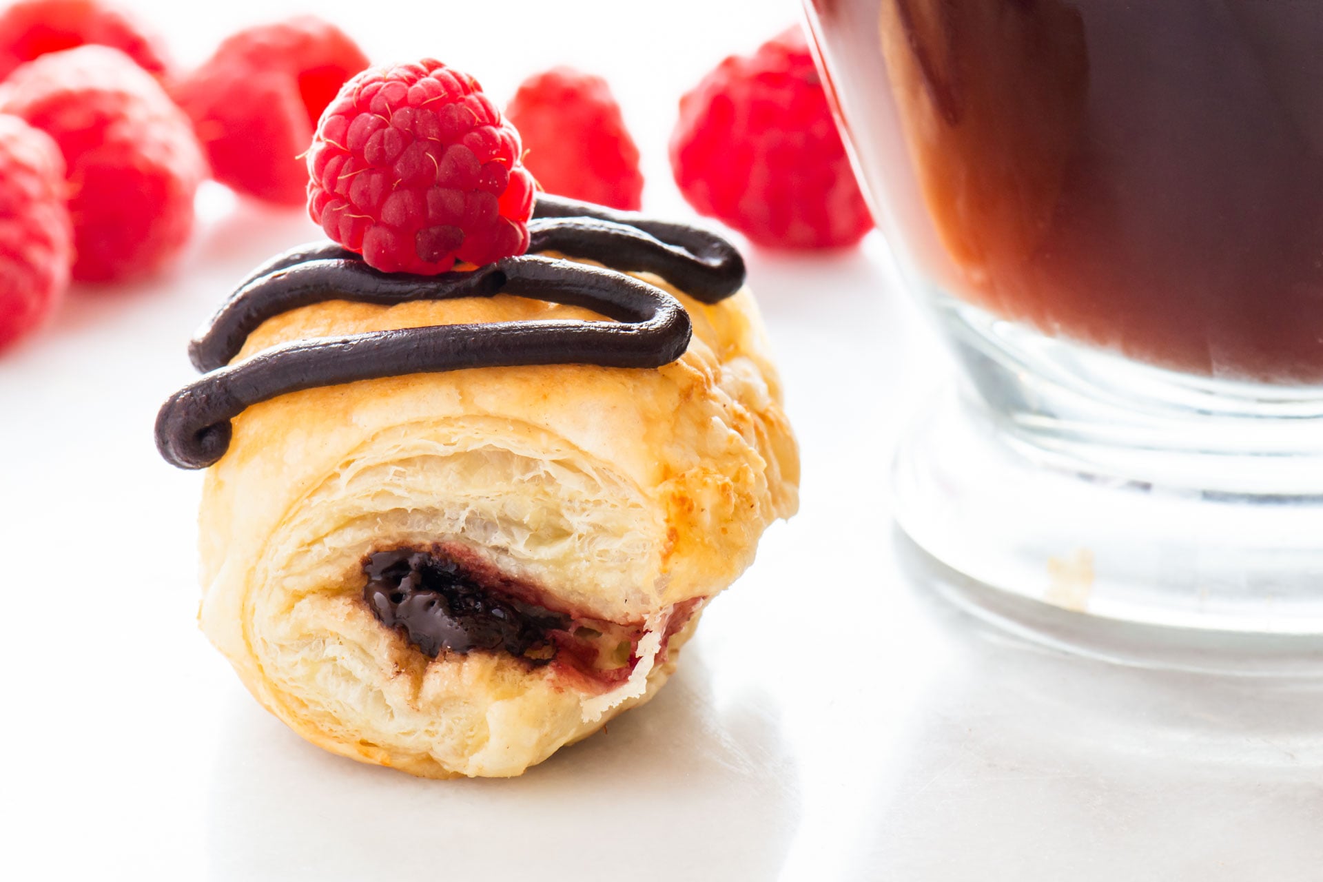 A Vegan Chocolate Croissants with chocolate drizzle and a red raspberry on top sits beside a glass mug of coffee.