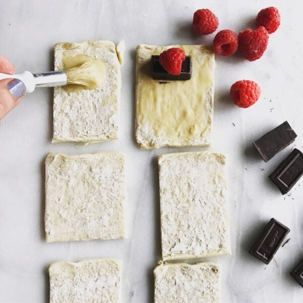 A hand reaches in to brush melted vegan butter on puff pastry sheets. Raspberries and chocolate pieces sit nearby.