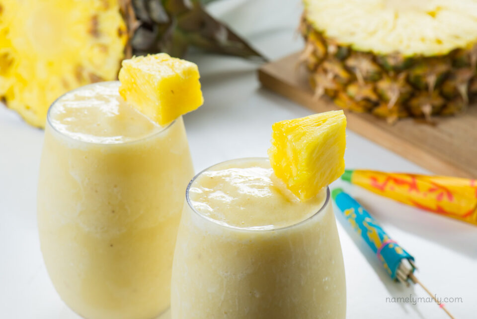 Vegan Skinny Pina Colada Cocktails sit beside each other with a fresh pineapple behind it and colorful umbrellas on the side.