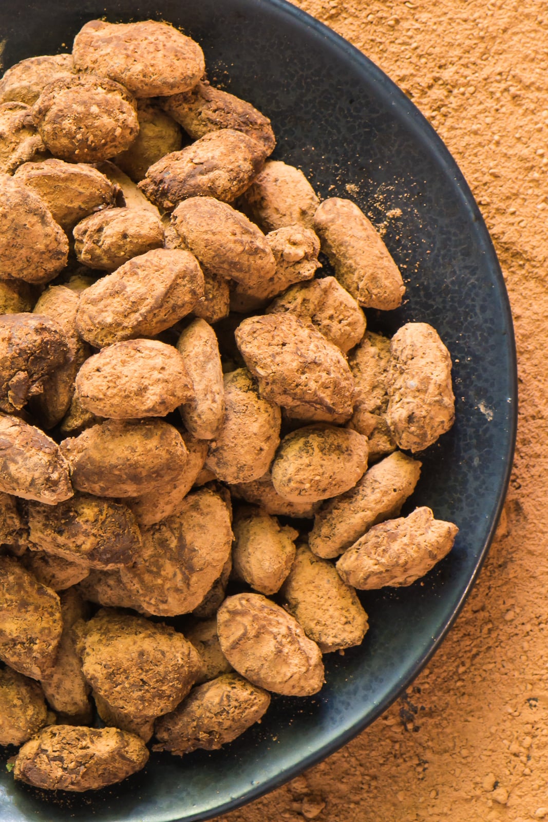 A plate full of dark chocolate covered almonds with cocoa powder sprinkled all around it.