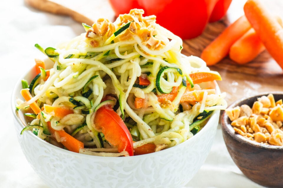 A bowl of zucchini noodles has chopped peanuts on top. There's a wooden bowl with more chopped peanuts on the side and a cutting board with veggies in the background.