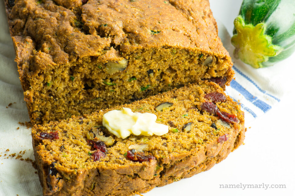 A loaf of zucchini bread has a slice cut out, sitting next to a zucchini. There's a pat of butter on the slice.