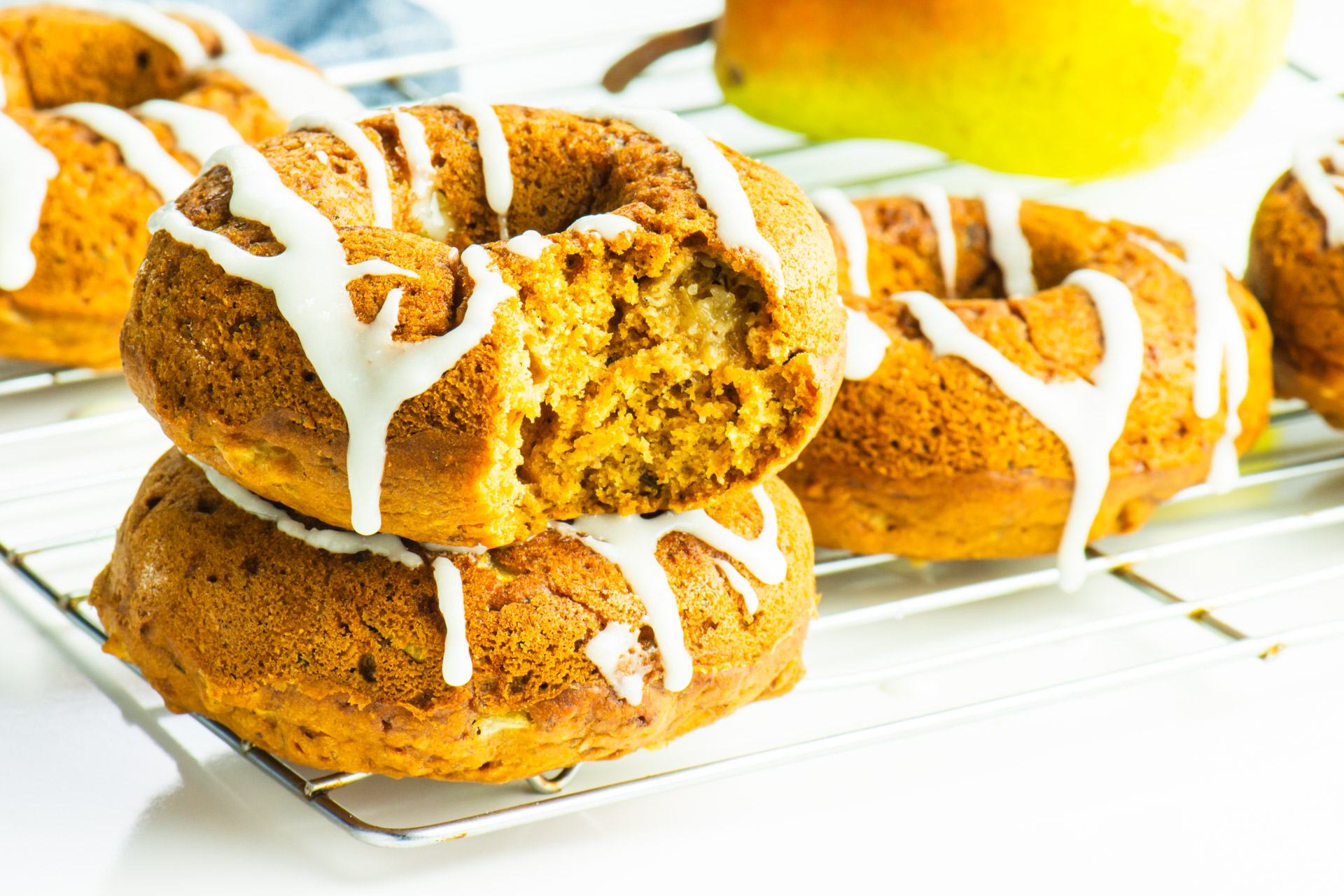 There are several ginger donuts stacked on a baking rack, one with a bite taken out. There's a pear and a blue kitchen towel in the background.

