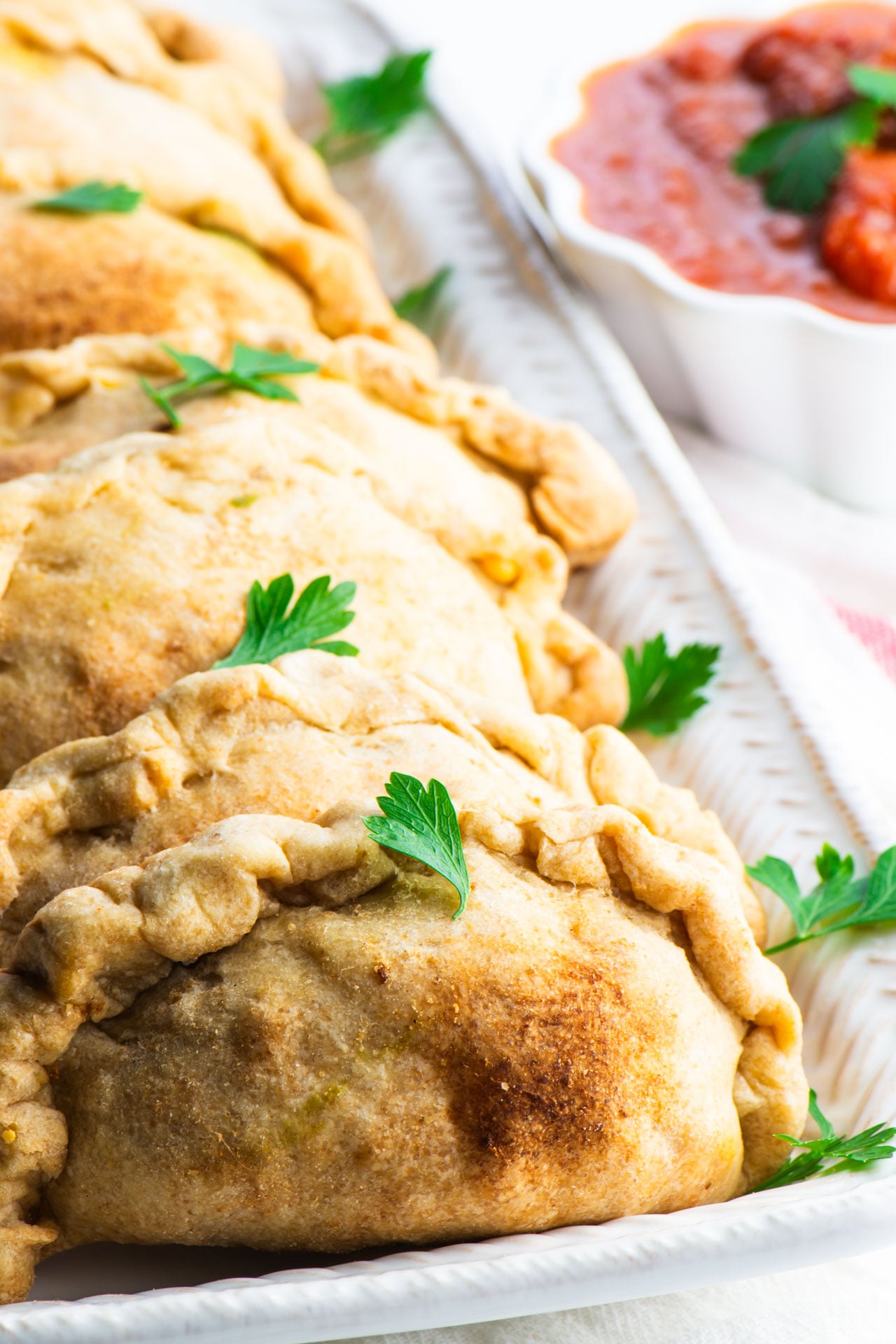 A row of  vegan beef empanadas on a serving platter next to a bowl of red sauce.