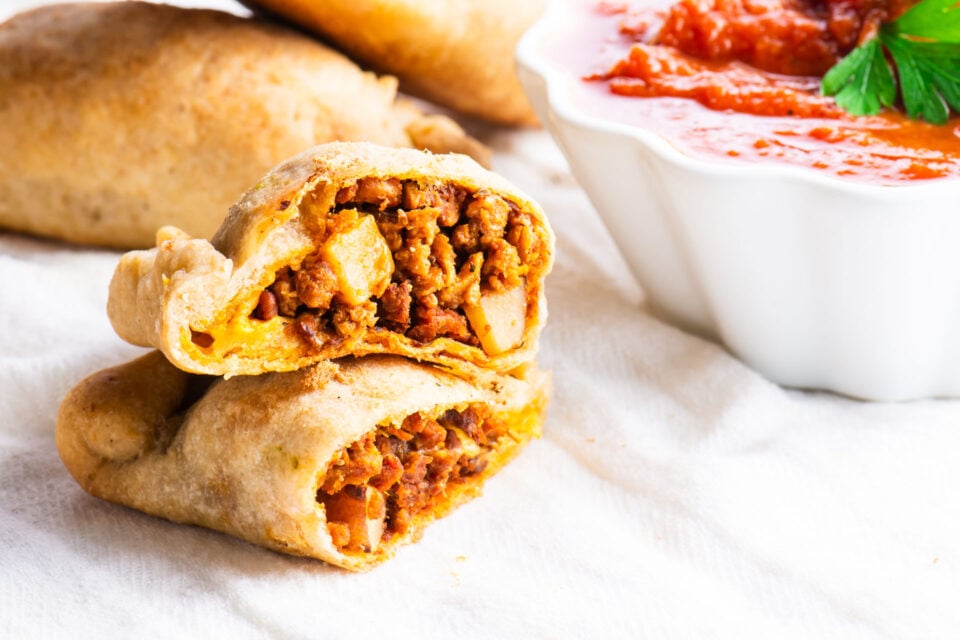 An empanada is cut and half and stacked. It sits next to red sauce and more empanadas behind it.
