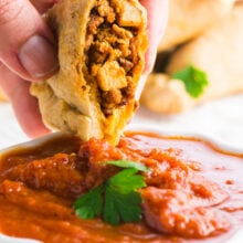A hand holds a vegan empanada over red sauce.