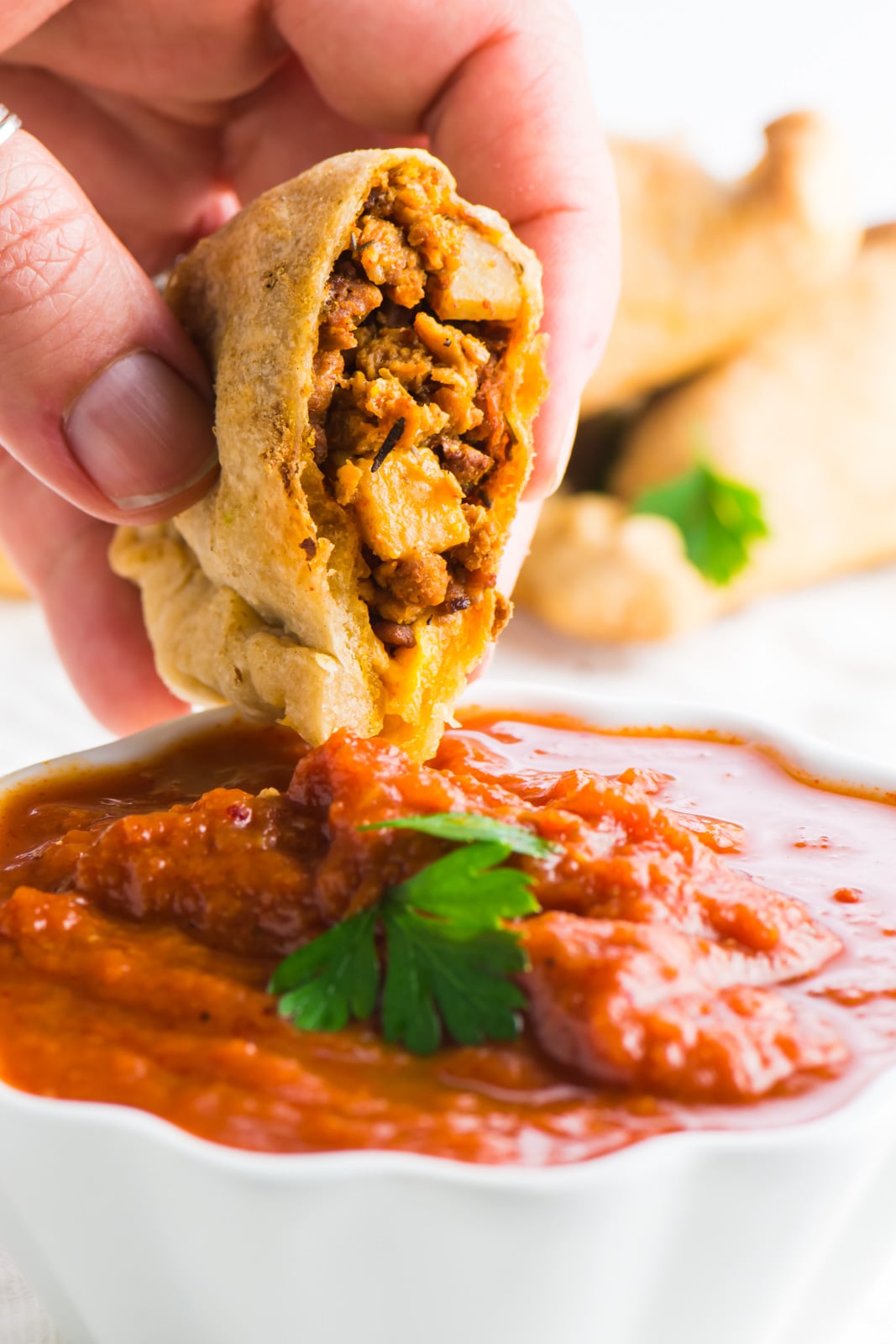 A hand holds a vegan empanada over red sauce.