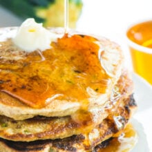 A stack of zucchini pancakes has maple syrup being drizzled over the top. More maple syrup and a zucchini is in the background.