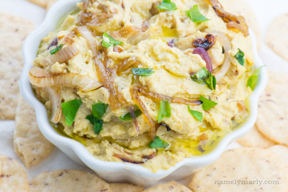 Vegan caramelized onion hummus I served in a small white bowl. There are crackers around it.
