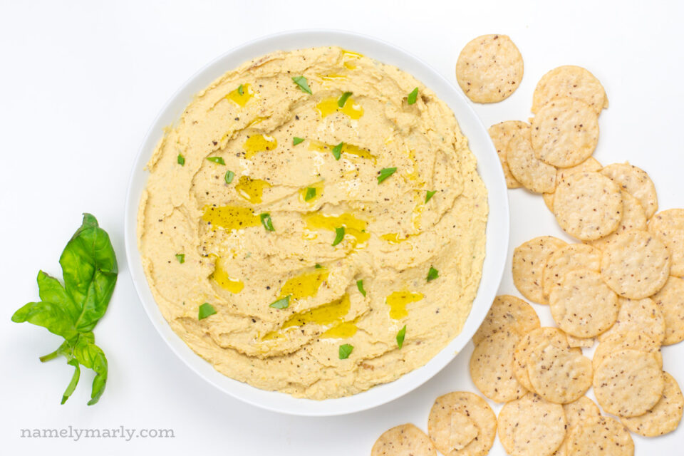Looking down on a bowl of hummus with chips beside it and fresh herbs on the other side.